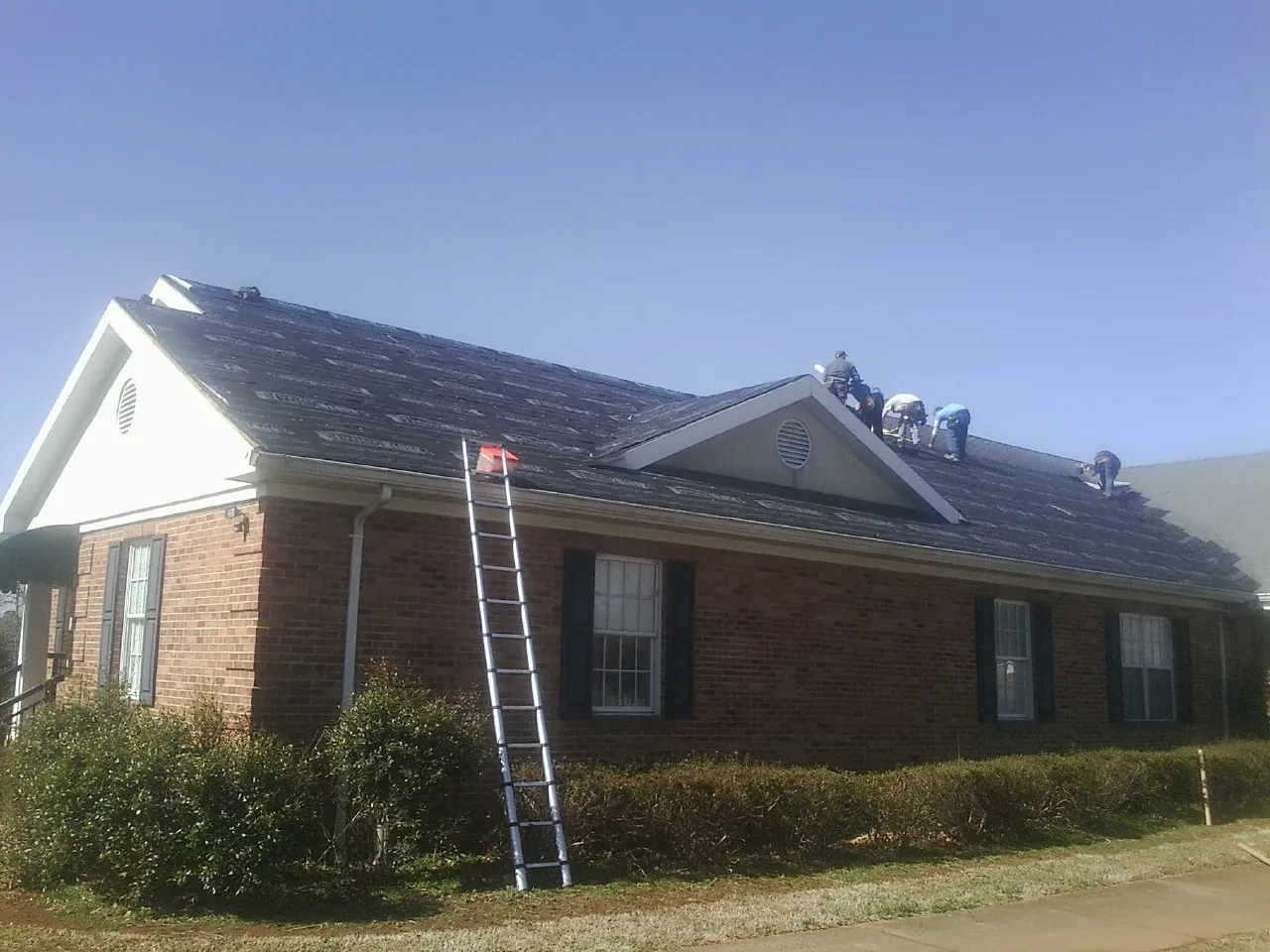 Crew tearing shingles off a brick ranch roof with a ladder set — Emergency Roof Repair work by our crew in Brenham